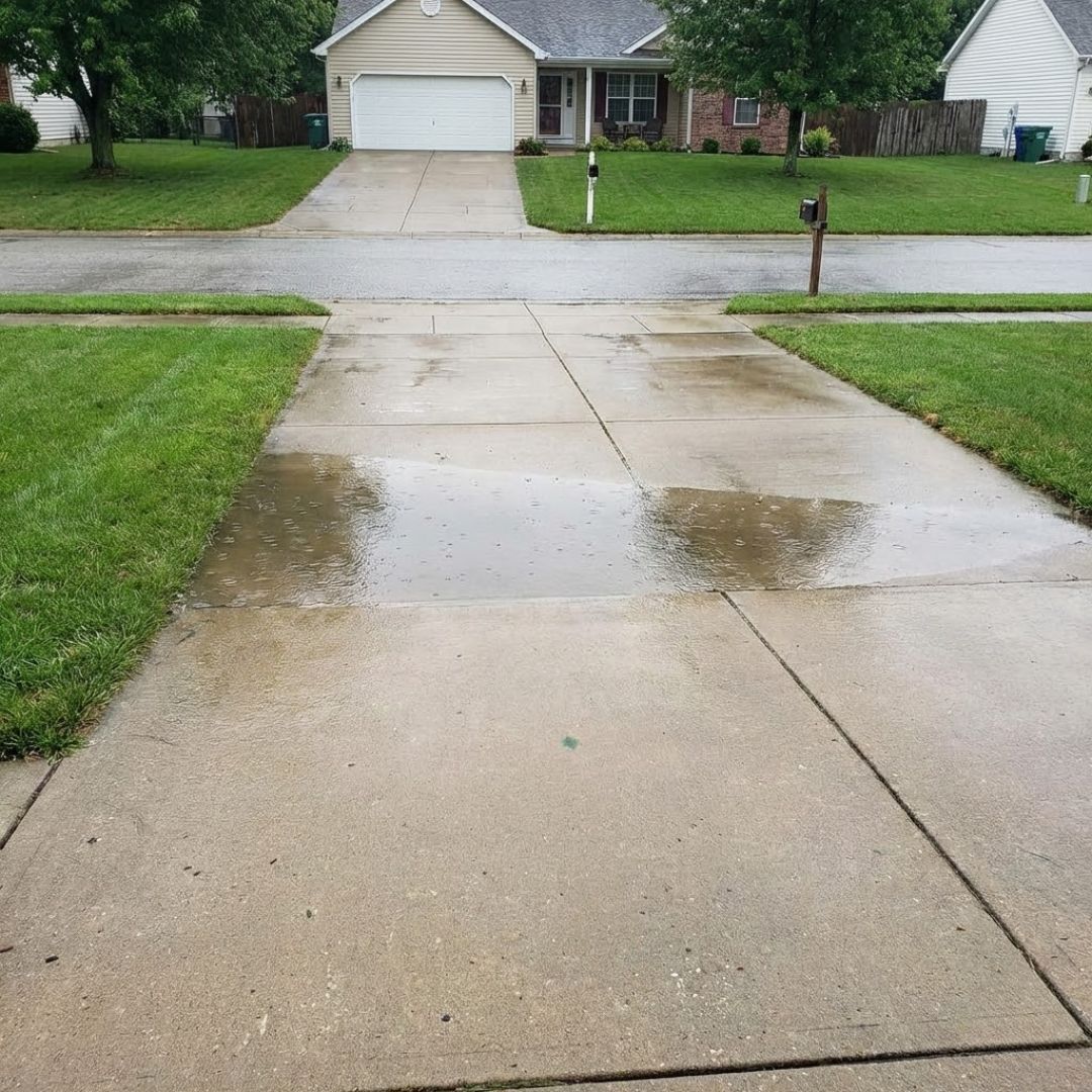 A residential concrete driveway with a large puddle of water from a recent rain.